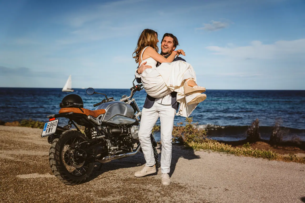 Groom carrying Antonia, laughing, beside a motorbike above the sea at La Escollera, Ibiza