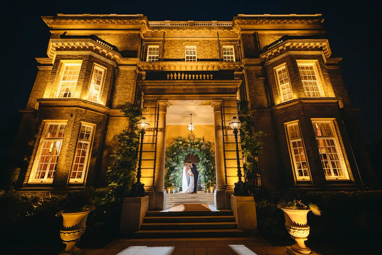 Ian and Jade kissing in the glowing floral-framed entrance of Hedsor House at night