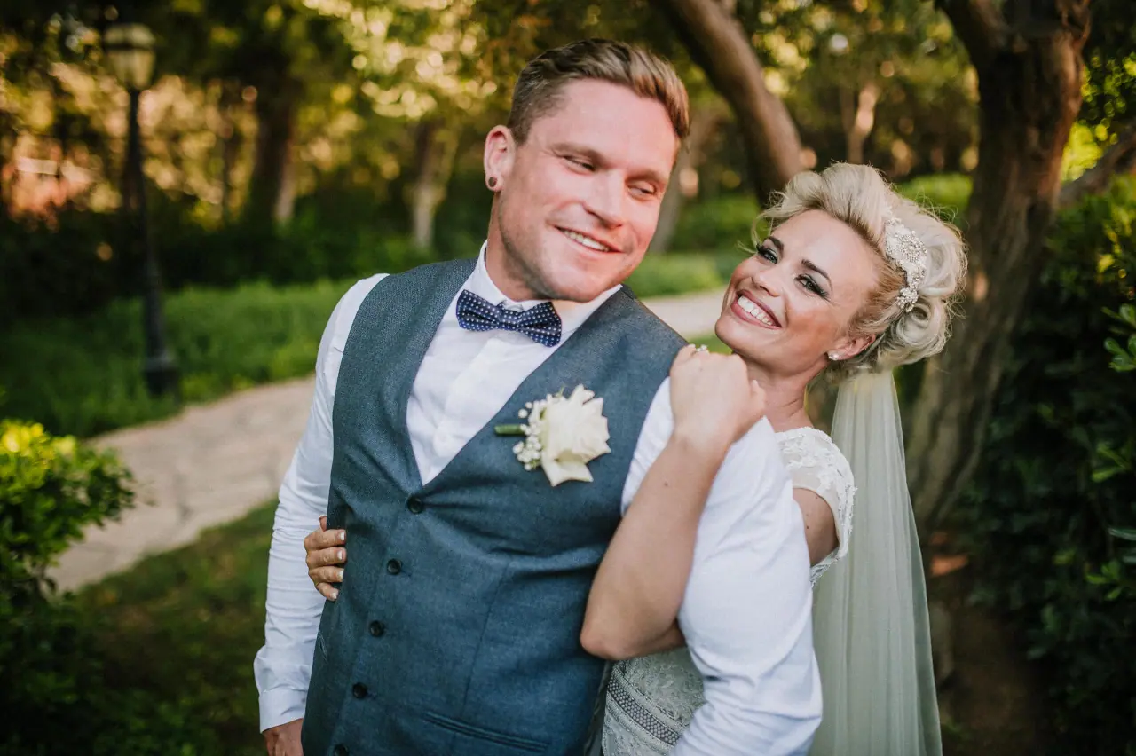 Smiling groom in a blue waistcoat as the laughing bride hugs him from behind in the gardens of Elysium, Paphos