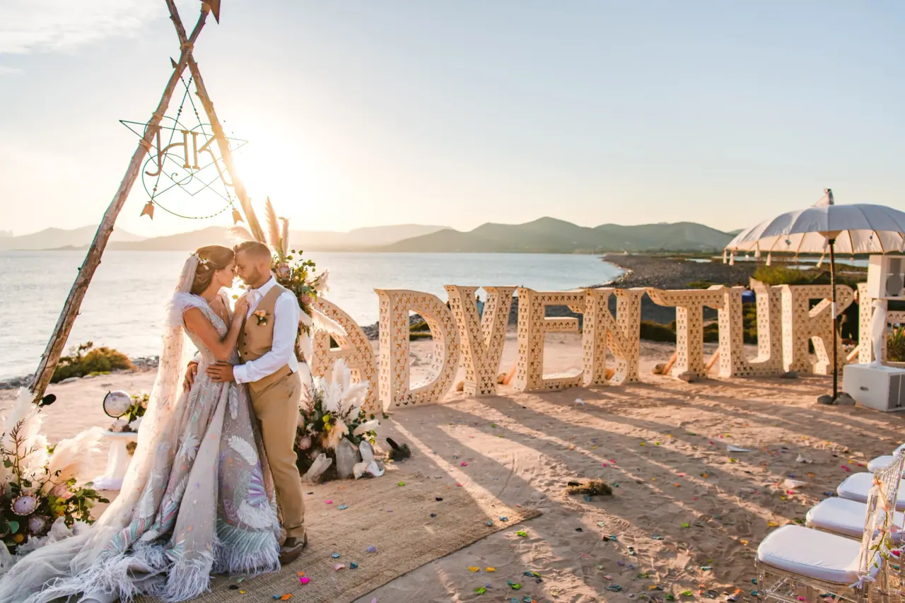 Kenton and Hassiba embracing beside giant ADVENTURE letters and a tipi arch at Experimental Beach, Ibiza