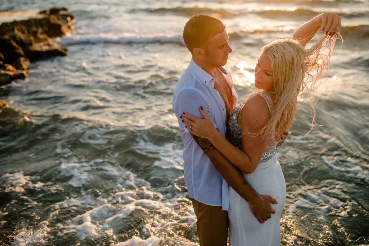 Maryellen and Ben embracing on sea rocks as waves wash around them in golden light, Paphos