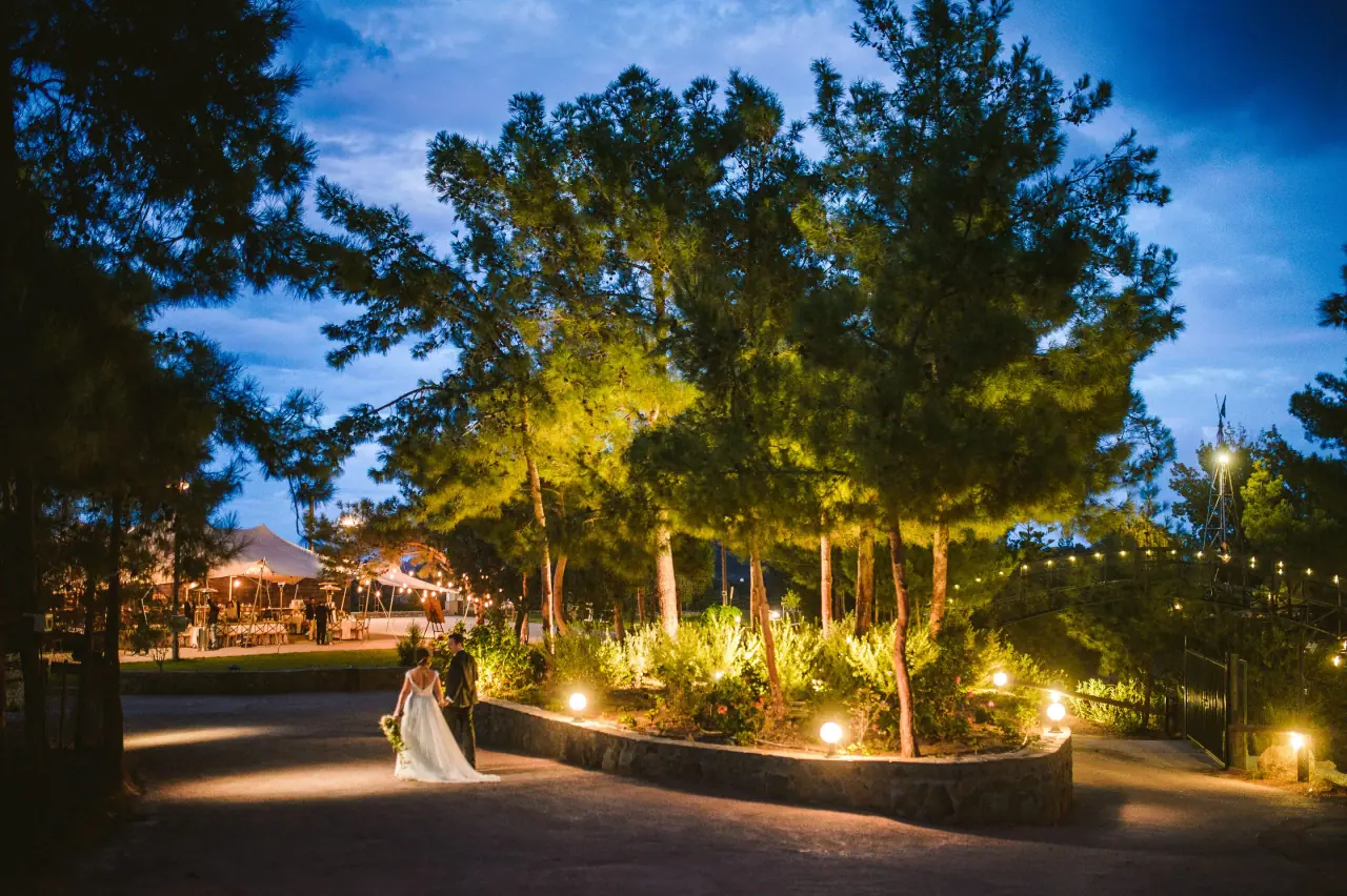 Nermine and Christopher walking a lantern-lit path under pine trees at dusk at Lexeko Estate, Cyprus
