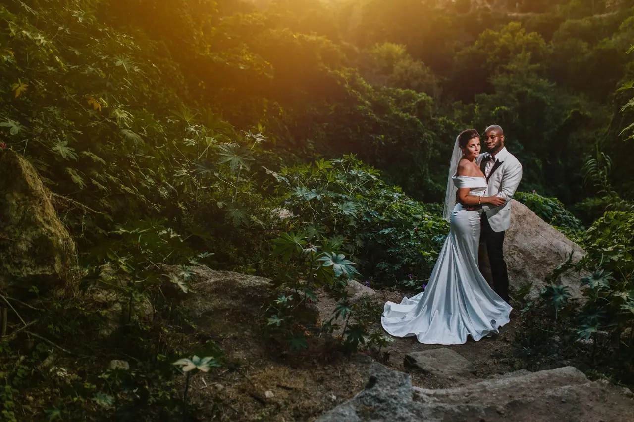 Reginald and Susannah embracing on rocks amid lush greenery in golden evening light, Paphos