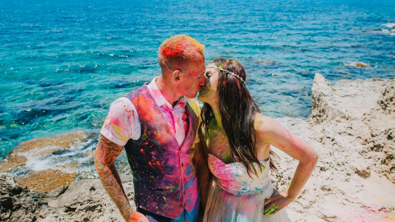 Sarah and Martin covered in colourful Holi powder, kissing on rocks above the turquoise sea at Nissi Beach, Ayia Napa