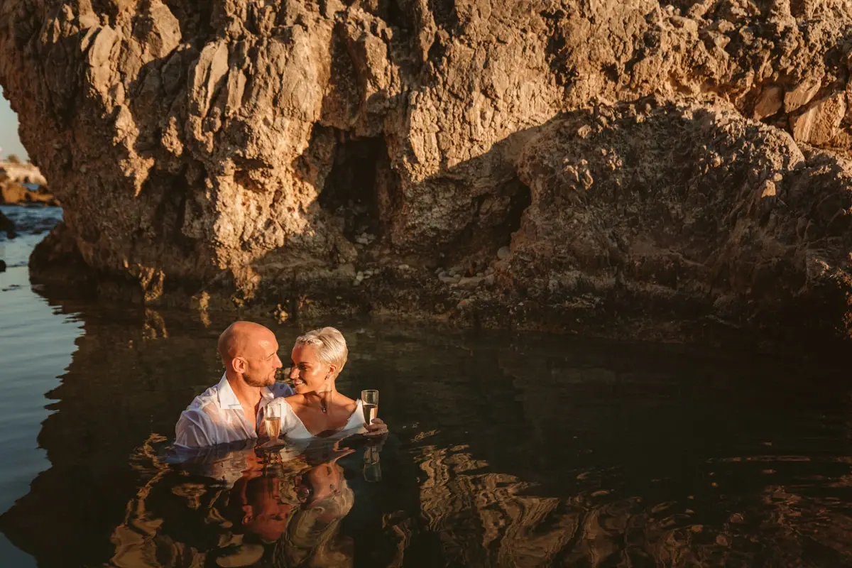 Alana and Luke sitting in a rocky sea pool in their wedding clothes, Paphos