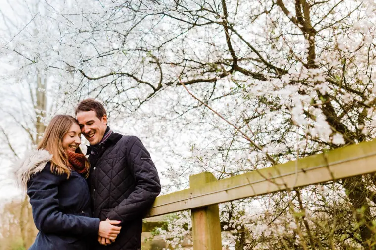 Couple cuddling beside a wooden fence under white spring blossom