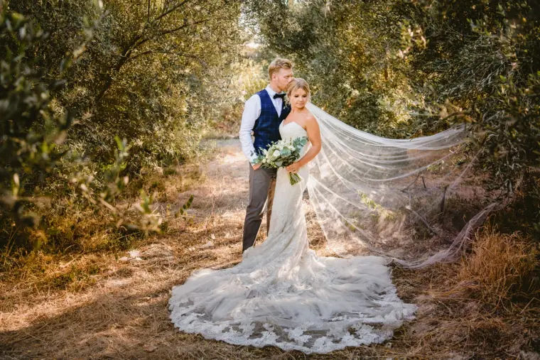 Fiona and Paul in an olive grove, her long lace veil and train spread across the path