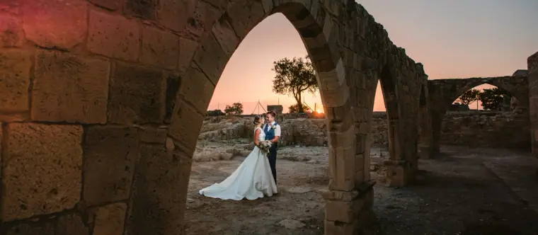 Bride and groom framed by an ancient stone arch at sunset, Kouklia