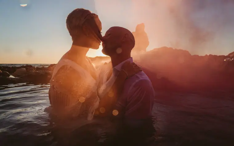 Alana and Luke kissing in the sea at sunset with pink smoke behind them