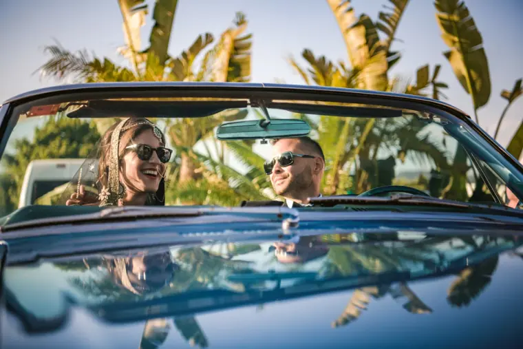 Kenton and Hassiba in sunglasses driving a classic convertible among palms, Ibiza