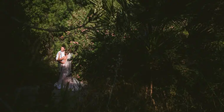 Amie and Graeme embracing in a patch of sunlight within dark, leafy gardens