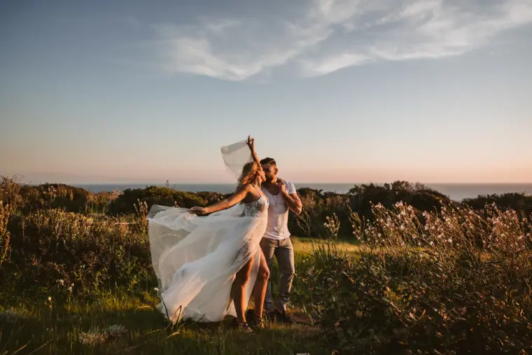 Bride twirling her dress in long grass by the sea as her partner looks on, Kouklia