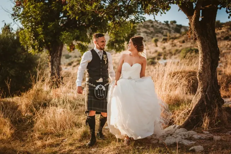 Hannah and Keith, in his kilt, walking through golden grass beneath trees at Vasilias Inn, Paphos
