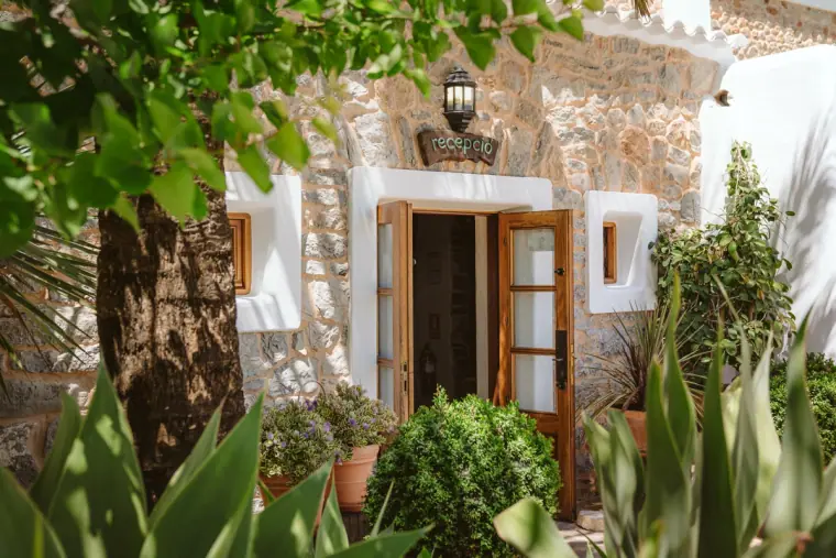 Stone farmhouse doorway framed by leafy trees and potted plants at Can Gall