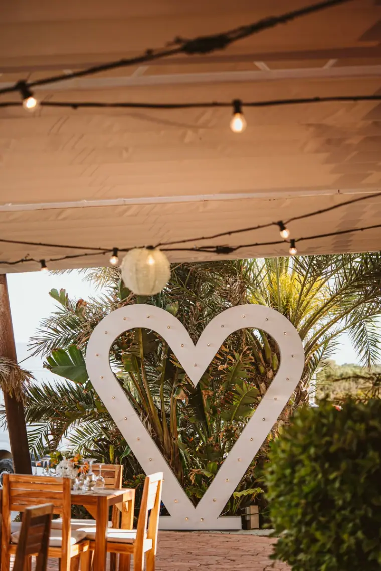 Large white heart sign beneath festoon lights among the palms on the restaurant terrace