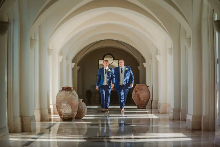 Groom and groomsman in navy suits walking through the arched corridor at Anassa Hotel