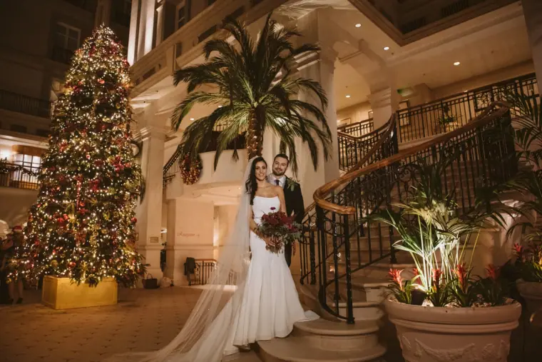 Bride with red bouquet beside a lit Christmas tree and palms in the lobby of The Landmark London