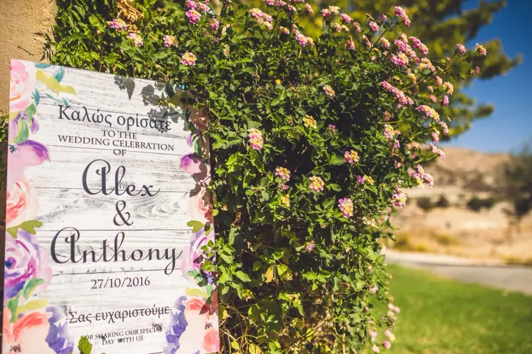Floral welcome board for Alex and Anthony's wedding in Greek and English beside a flowering shrub