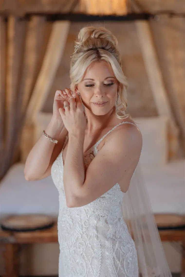 Bride fastening her earring in a lace gown, a canopy bed behind her