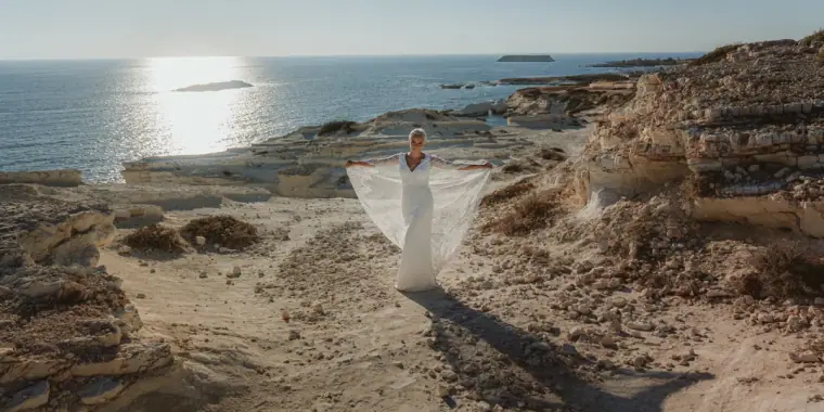 Alana in a flowing white dress walking across rocky coastline towards the sea