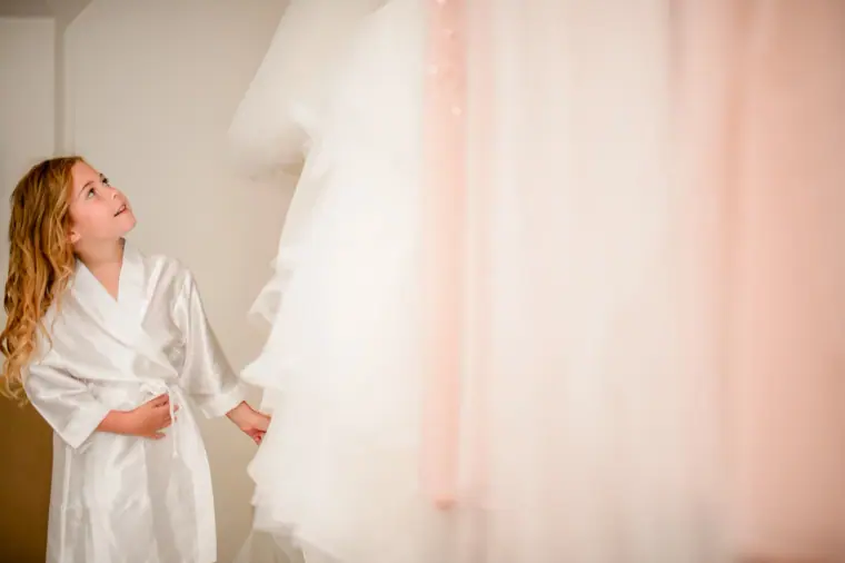Flower girl in a white robe gazing up at the bridal gown hanging by the window