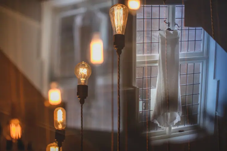 Wedding dress hanging in a leaded window beside glowing Edison bulbs