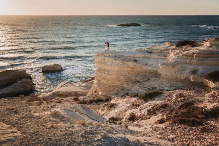 Aiko and Erez, tiny on a white clifftop above the sea at sunset, Sea Caves, Paphos