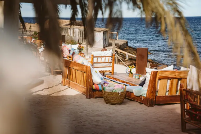 Beach lounge with wooden sofas and cushions framed by palm leaves by the sea at La Escollera
