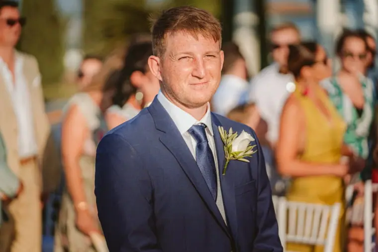 Jack in a navy suit waiting for his bride as guests gather at Anassa Hotel
