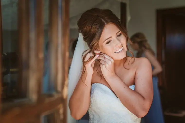 Hannah smiling as she puts on her earrings by a wooden window