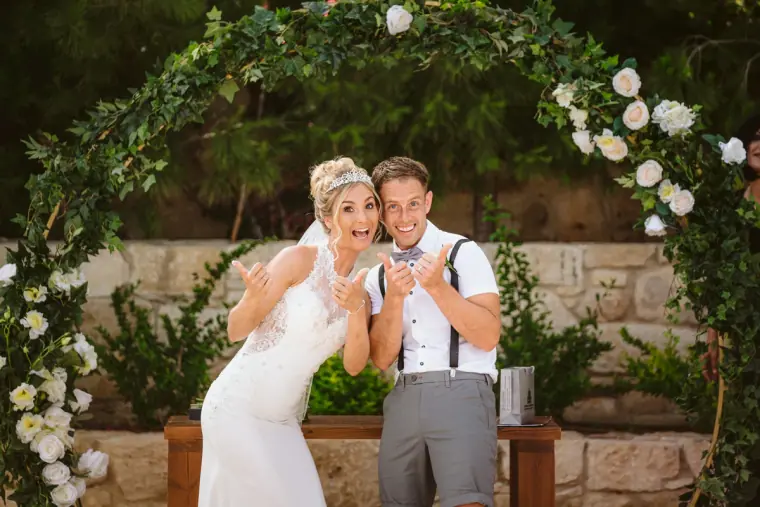 Victoria and James pulling playful faces beneath a floral arch at Liopetro, Kouklia