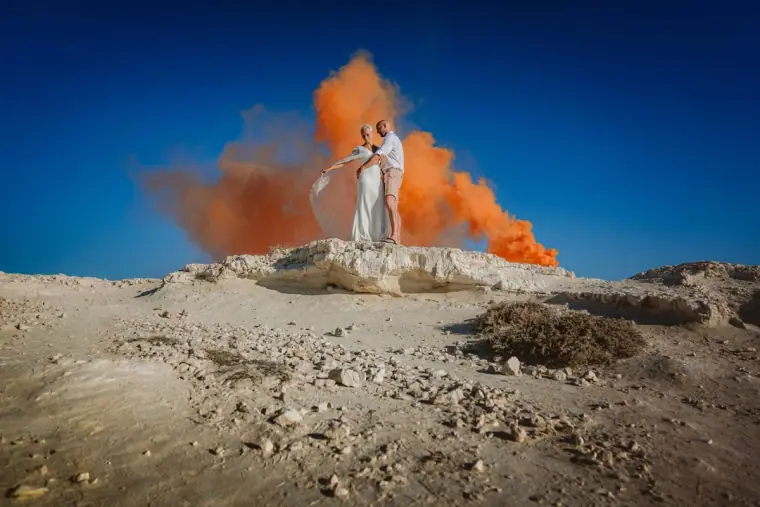 Alana and Luke on a sandy ridge with orange smoke against a blue sky, Paphos