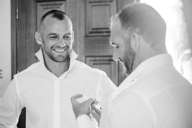 Black-and-white photo of the groom laughing as a friend fastens his cufflinks