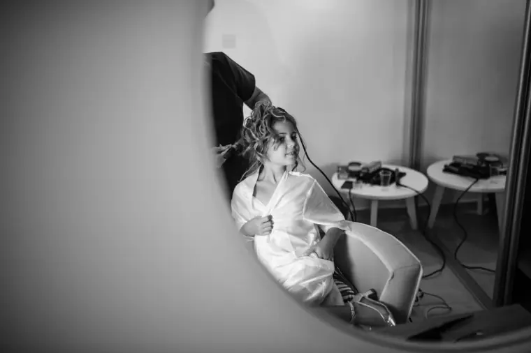 Black-and-white photo of the bride in her robe having her hair styled, seen through a doorway