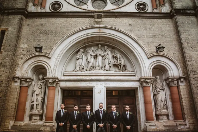 Groom and groomsmen in dark suits lined up beneath the carved stone church entrance