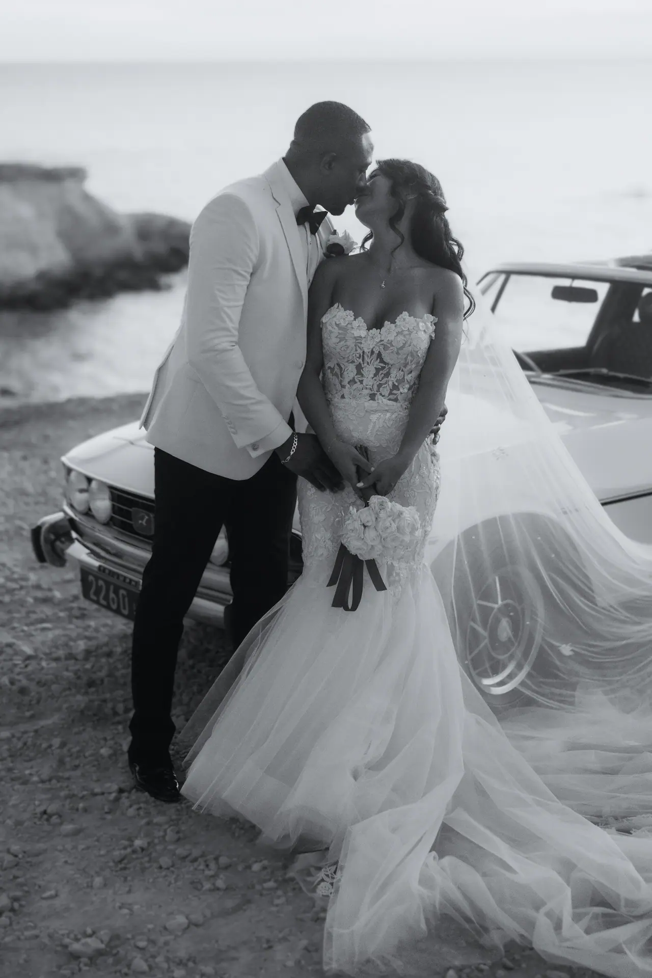 Black-and-white photo of the couple kissing beside the Triumph Stag on the Cyprus coast