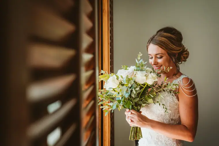 Bride smelling her white rose bouquet by a shuttered window
