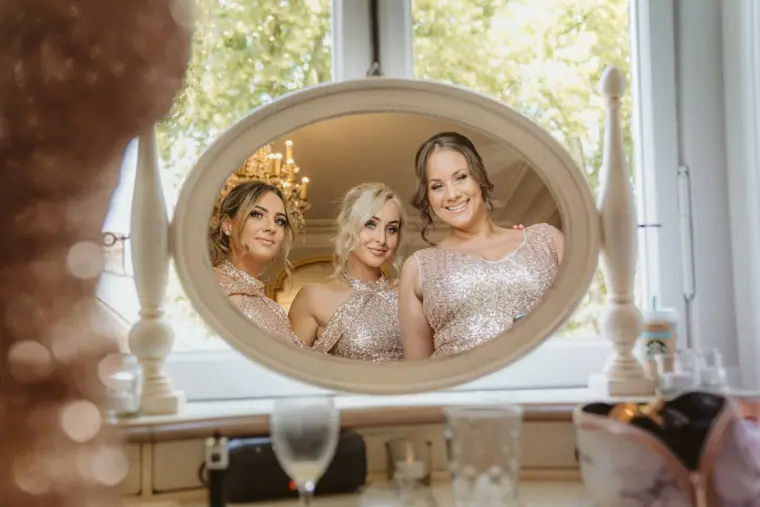 Bridesmaids in blush dresses reflected in an oval mirror while getting ready