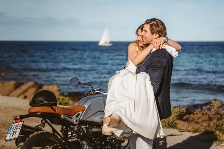 Groom lifting the bride in a hug beside a motorbike with a sailing boat on the sea behind