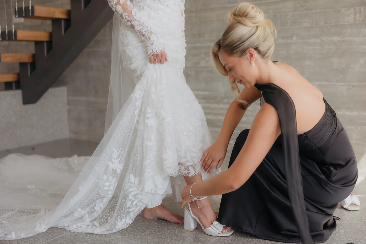 Bridesmaid in a black dress fastening the bride's shoe beneath her lace gown