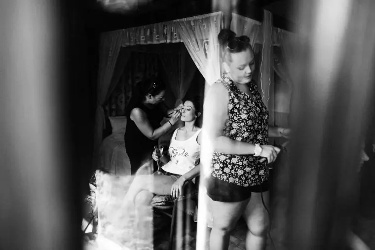 Black-and-white photo of the bride having her make-up done beside a canopy bed