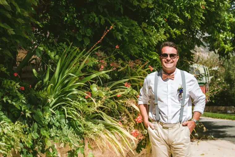 Graeme smiling in sunglasses, braces and a buttonhole among garden greenery