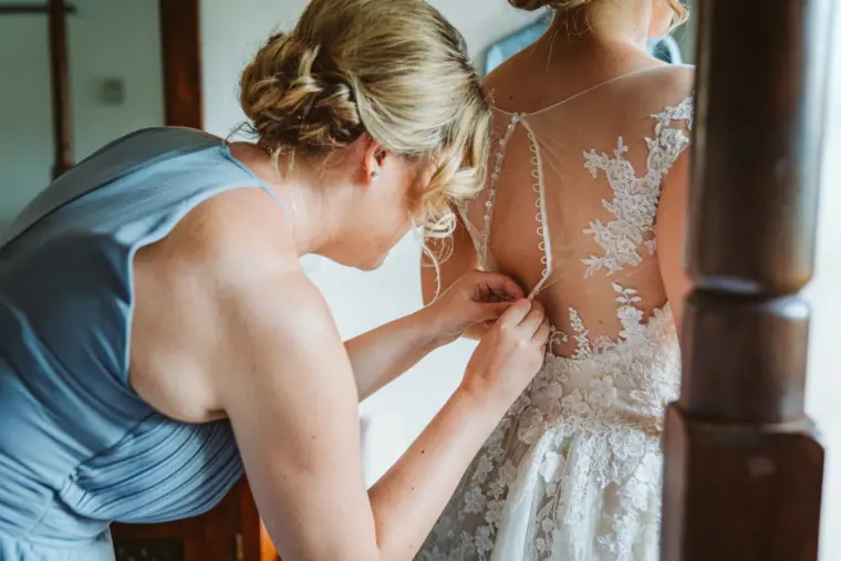 Bridesmaid in dusty blue buttoning the back of the bride's lace dress