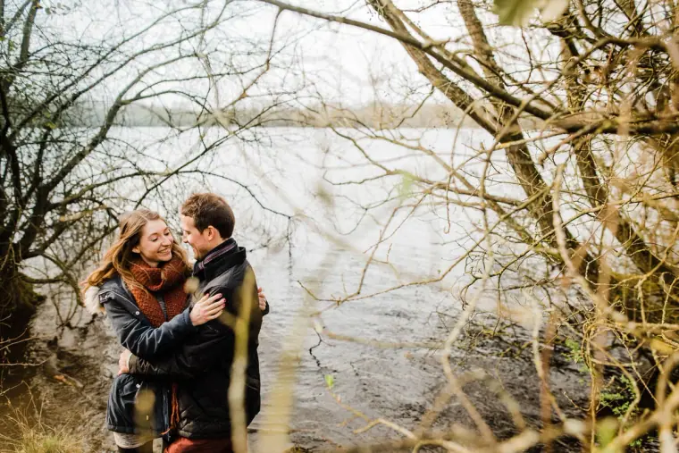 Couple hugging at the water's edge, framed by bare branches