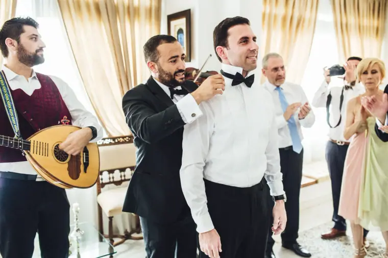 Angelos in a tuxedo being dressed by family as musicians play