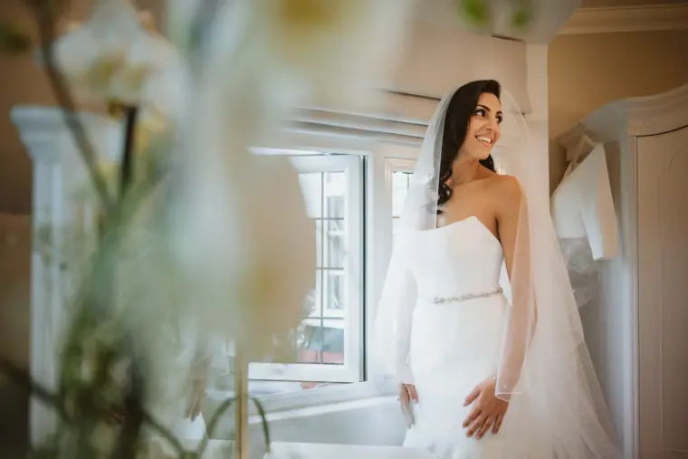 Bride in a strapless gown and veil standing by a window in soft light