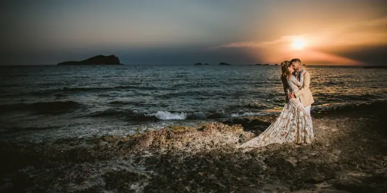 Kenton and Hassiba embracing on the rocky shore as the sun sets over the sea, Ibiza