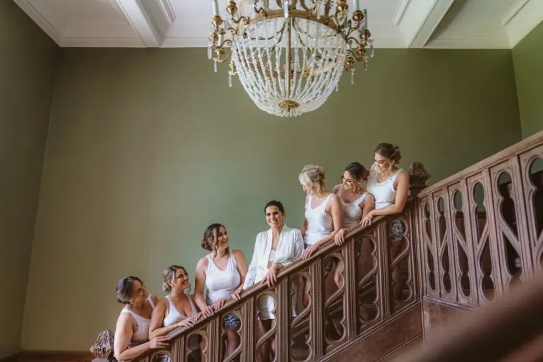 Bridesmaids in white robes lined up on a château staircase beneath a crystal chandelier