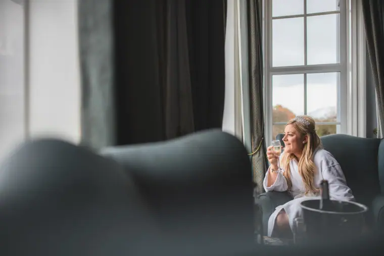 Bride in a robe sipping a drink in an armchair by the window