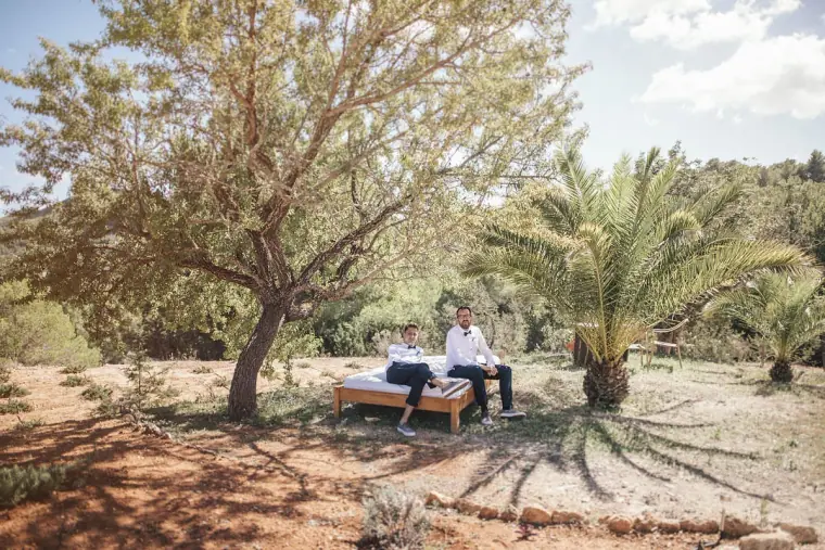 Groom sitting with a friend on a daybed beneath a tree and palm